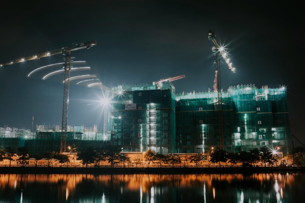 Night view of a construction site with cranes and office buildings reflected in a river.